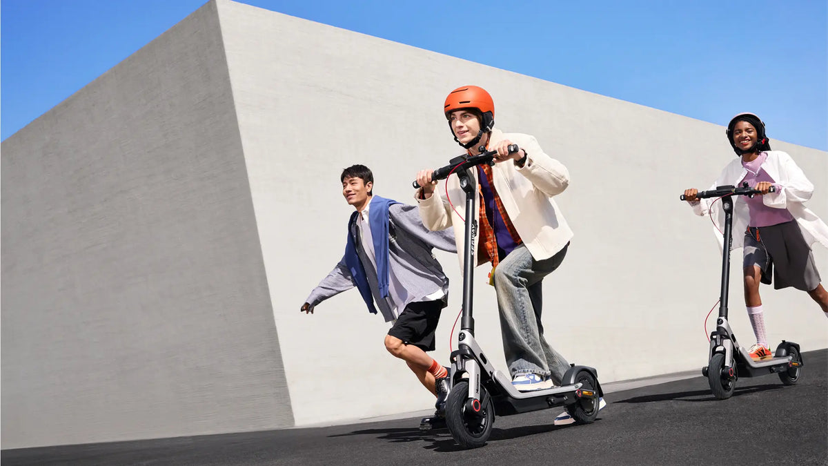 Trois jeunes sur des trottinettes électriques Ninebot E3, portant des casques, devant un mur blanc sous ciel bleu.